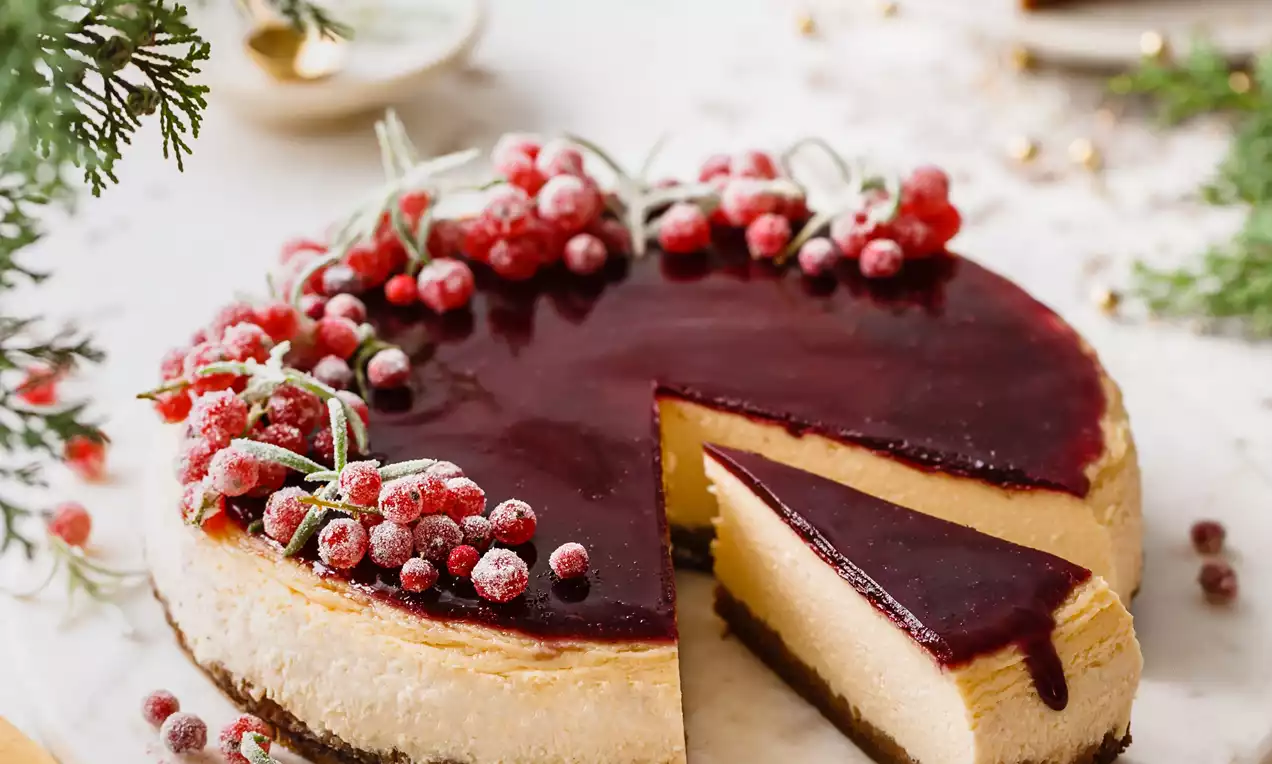 Picture - Creamy cheesecake with a biscuit base, glossy deep red topping, sugared berries and frosted rosemary sprigs on a white plate