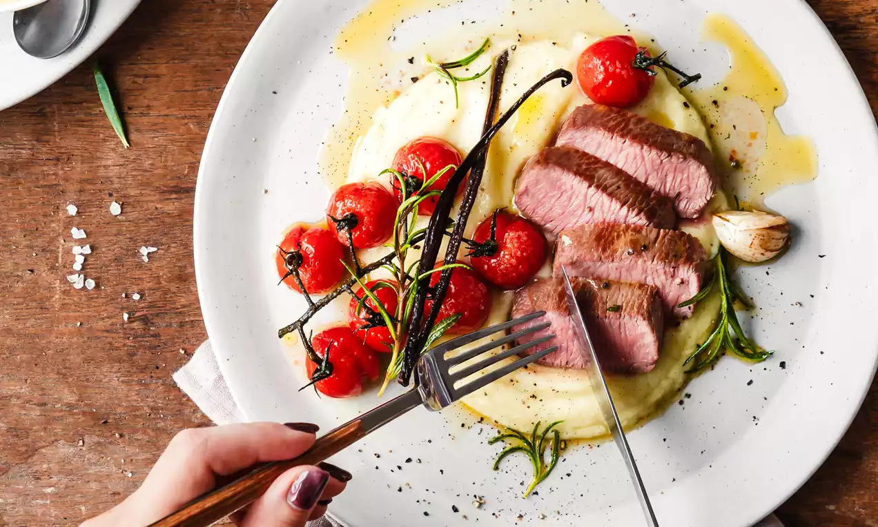 Picture - Slices of browned lamb loin on creamy vanilla-potato-puree next to glossy tomato confit with herbs on a white plate