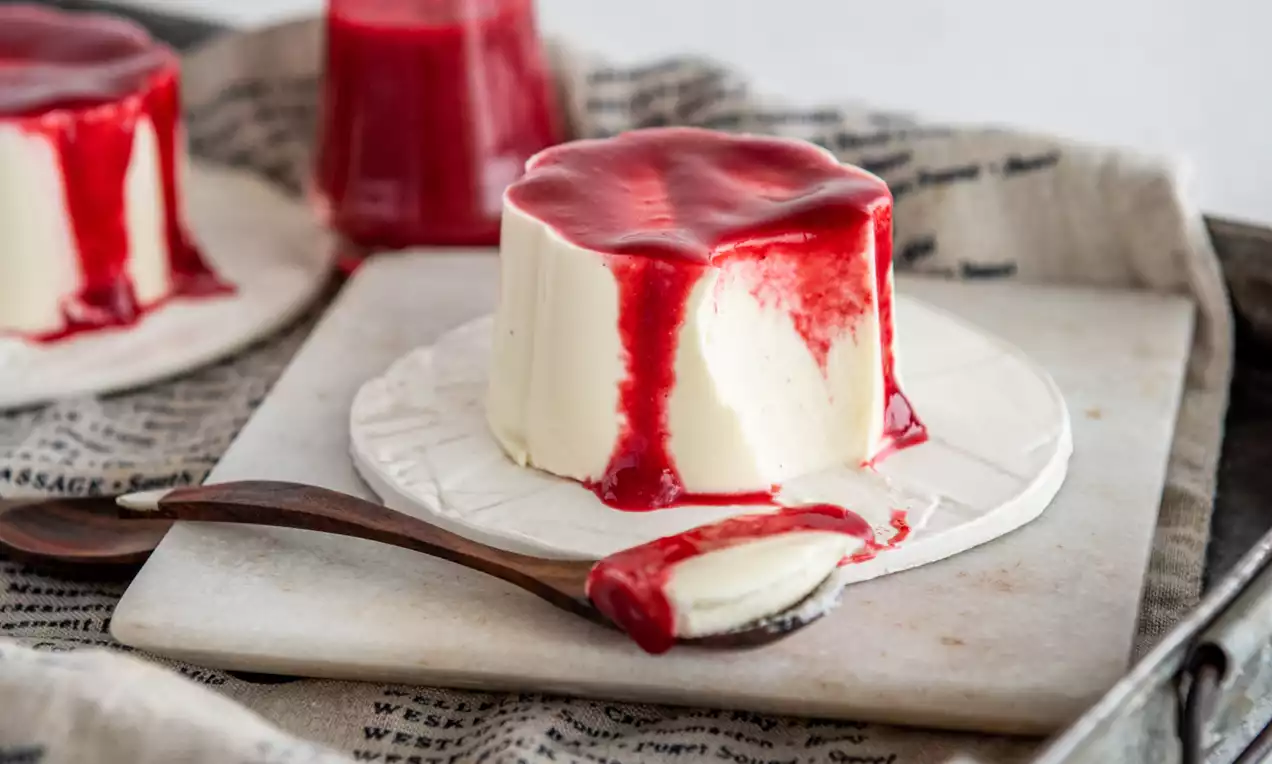 Picture - Silky, ivory Vanilla Panna Cotta draped with glossy red berry sauce, set on a white plate atop rustic paper in a metal tray