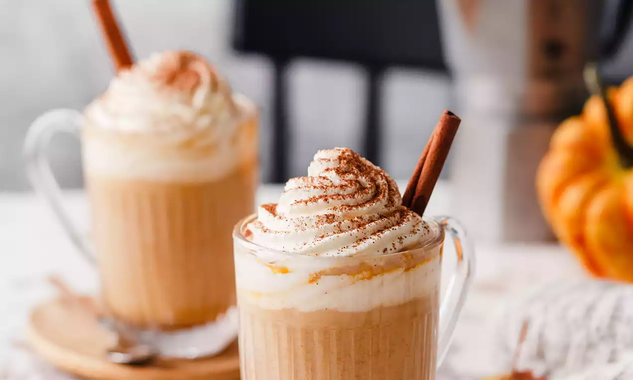 Picture - Creamy pumpkin spice vanilla latte in a clear glass with swirled whipped cream, dusted with cinnamon, garnished with a cinnamon stick