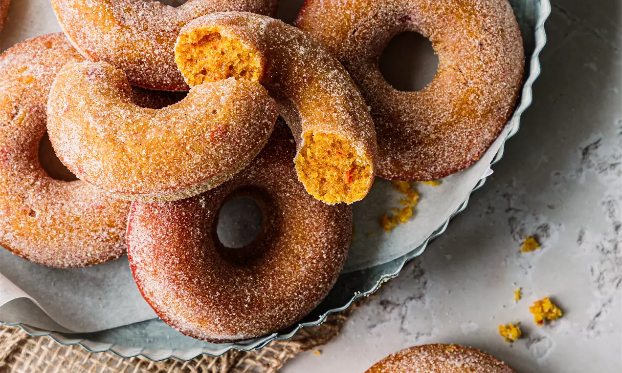 Picture - Pumpkin doughnuts golden-orange with a sugared coating, some whole and one bitten, arranged on a blue plate with burlap and crumbs
