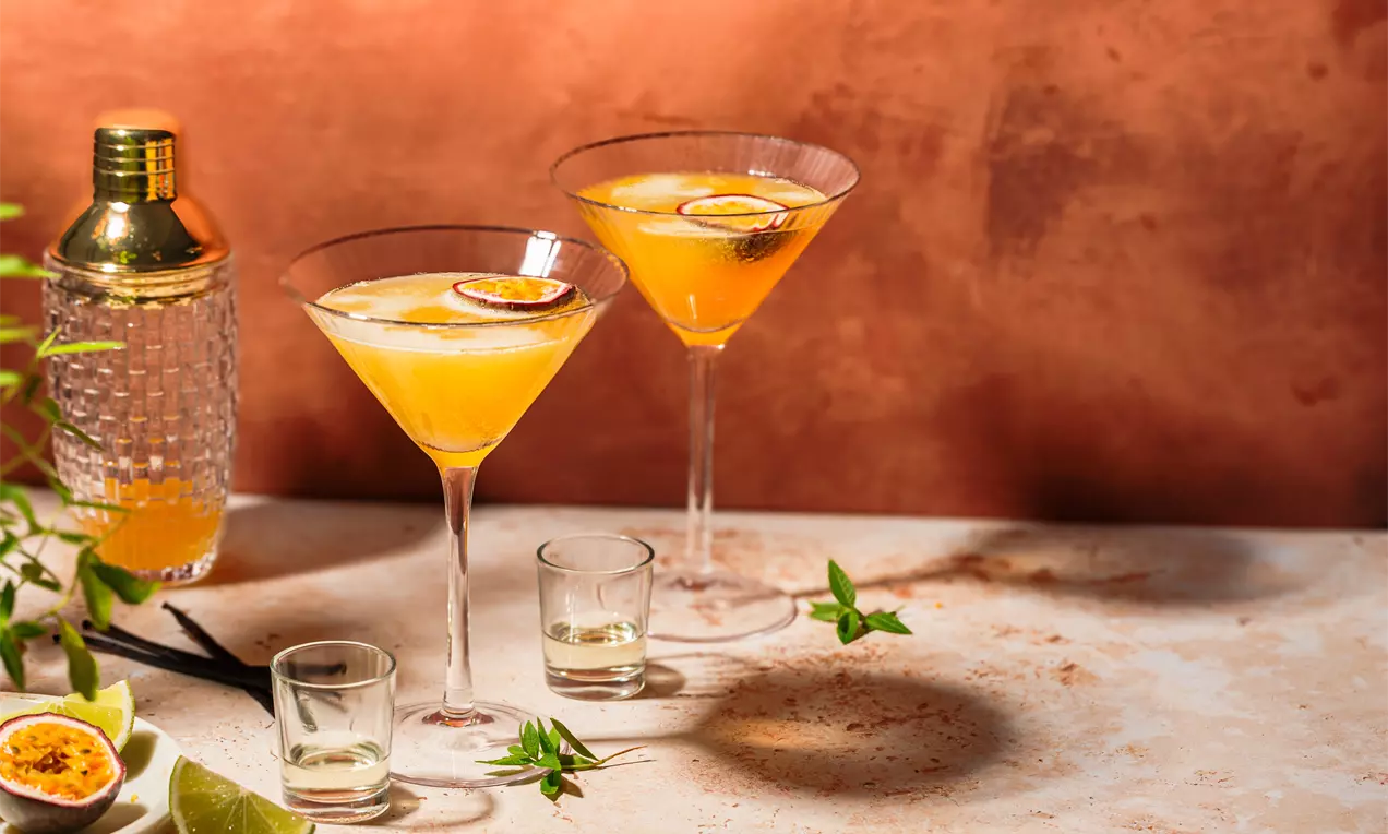 Picture - Two elegant martini glasses with bright amber-orange cocktails, topped with half passion fruits, on a textured surface with fresh limes