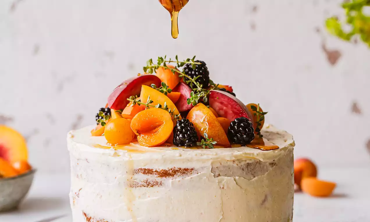 Picture - Vanilla sponge cake with rustic white frosting, topped with vibrant fresh fruits and herbs, drizzled with golden honey on a white plate
