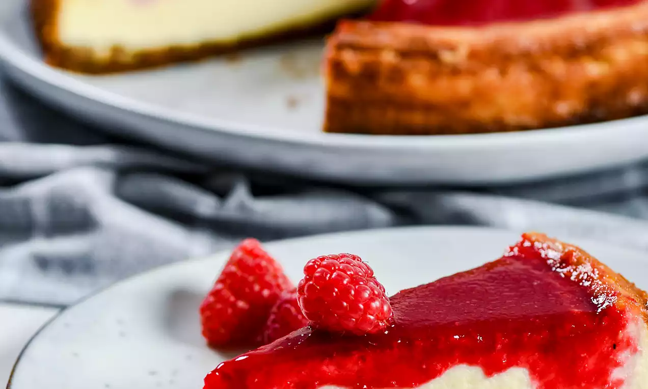 Picture - Baked raspberry cheesecake with vivid red topping, creamy filling on a biscuit base, garnished with fresh raspberries on a white plate