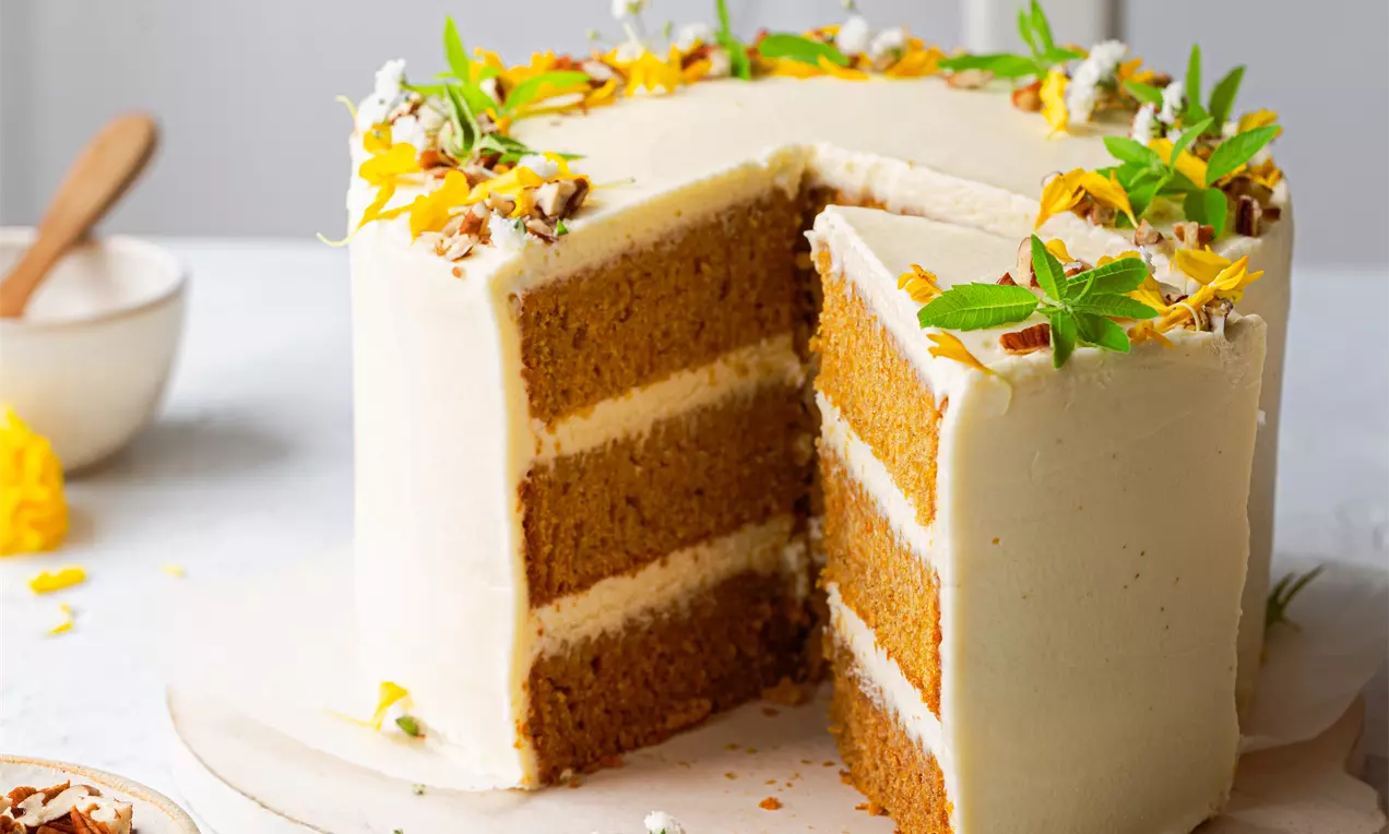 Picture - Carrot cake with three moist orange layers frosted in smooth white icing, topped with yellow petals, nuts, and fresh green sprigs