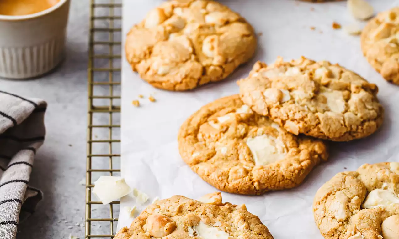 Picture - Golden round cookies with a crumbly texture and chunks of white chocolate and macadamia arranged on parchment on a cooling rack