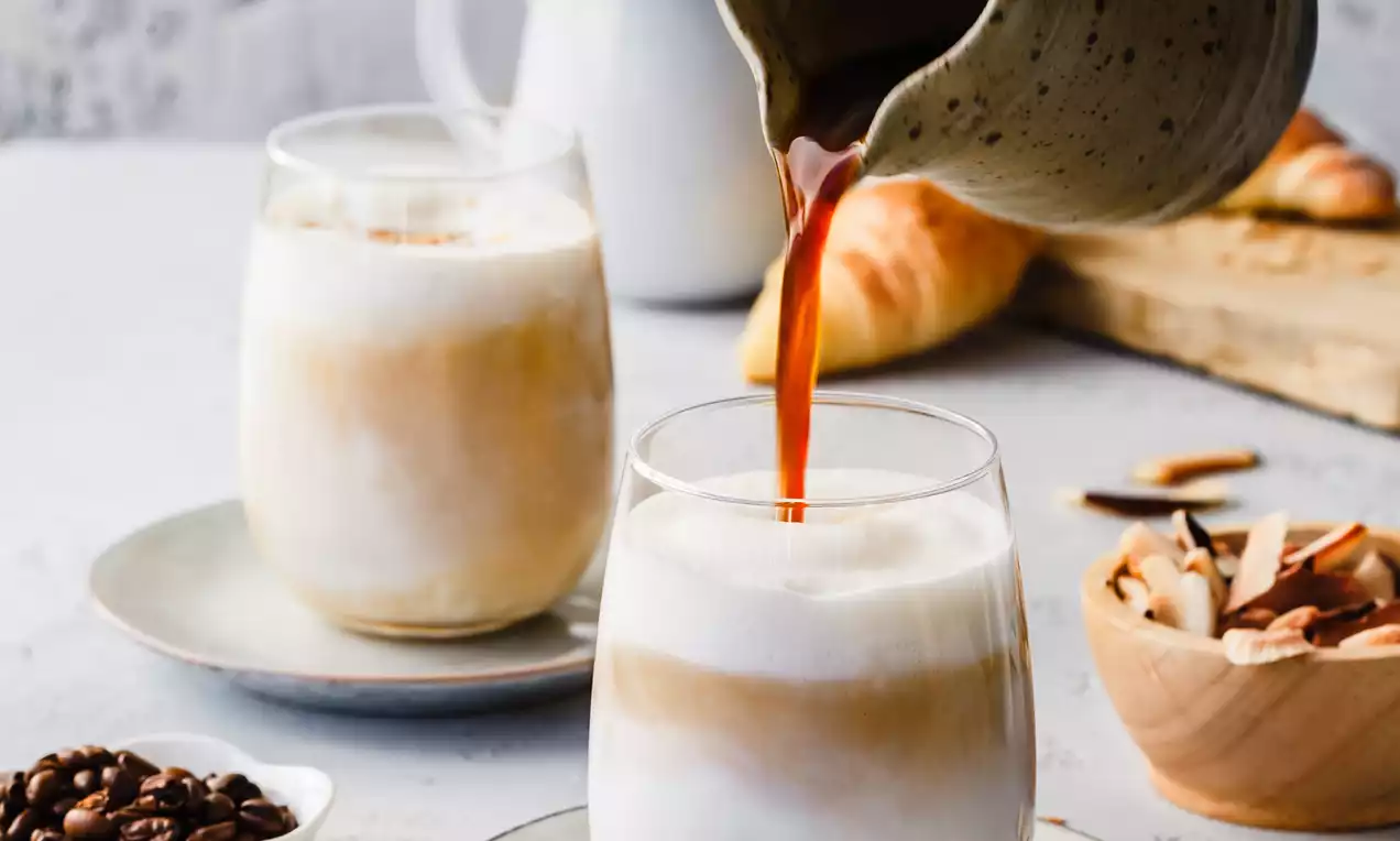 Picture - Layered coconut vanilla latte in a clear glass with swirls of white and caramel, served on a saucer amidst scattered coffee beans and almond slices