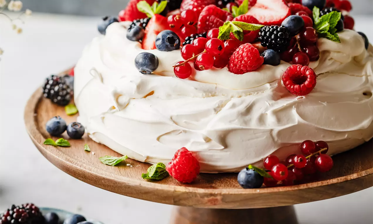 Picture - White vanilla pavlova with glossy swirls, topped with vibrant berries and fresh mint, arranged on a round wooden cake stand